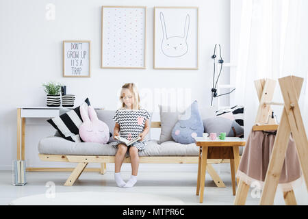 Cute young girl reading book on stylish sofa in white room Stock Photo