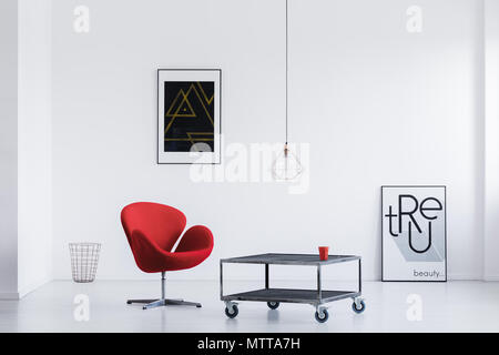 Stylish waiting room with white walls, industrial table, red armchair and modern posters Stock Photo