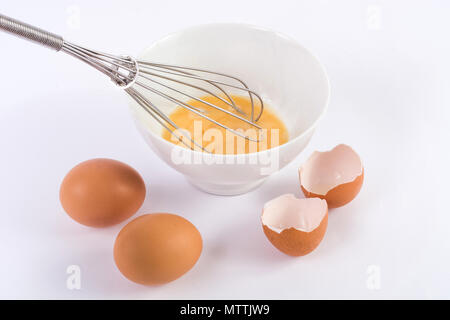 Whipped eggs in bowl with whisk and two egg and one broken on kitchen white table. Cooking and food concept Stock Photo
