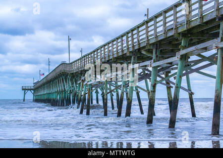 Under the Pier - beautiful ocean views from Oak Island beach, crashing waves, and the wooden pier on a cloudy day. Stock Photo