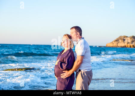 Portrait of happy love pregnant couple embracing each other and watching ahead to the sea during walk on the beach. Relax by the calm sea in sunshine. Stock Photo