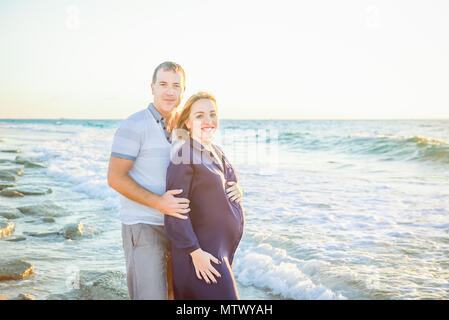 Portrait of happy love pregnant couple embracing each other and watching to the sea during walk on beach. Relax by the calm sea in sunshine. Family va Stock Photo