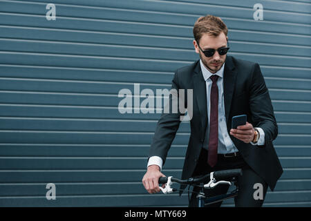 Young businessman in sunglasses using smartphone while riding bicycle Stock Photo