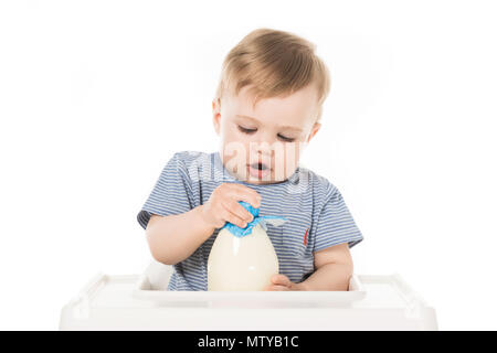 little boy trying to open bottle of milk and sitting in highchair isolated on white background Stock Photo