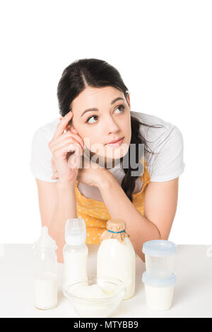 attractive woman in apron with finger on temple at table with plastic containers, bowl, bottle of milk and baby bottles Stock Photo