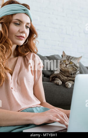 beautiful young woman working with laptop while sitting near her tabby cat at home Stock Photo