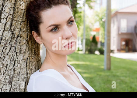 close-up shot of thoughtful young woman leaning back on tree trunk in park and looking away Stock Photo