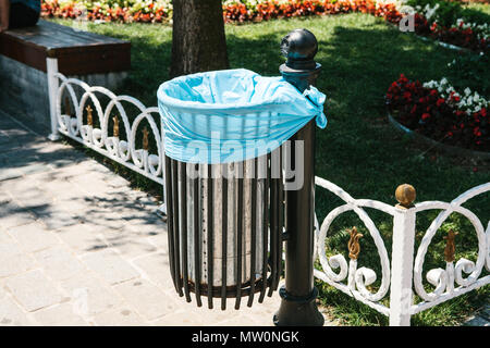 Close-up - iron trash urn with one-time package inside on the background of benches, fences and flowers in sunny park Stock Photo