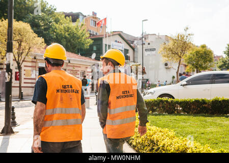 Two unknown men in work clothes - protective helmets and yellow waistcoats are walking along the sunny city Stock Photo