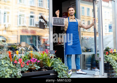 handsome young male florist in apron holding open sign and smiling at camera