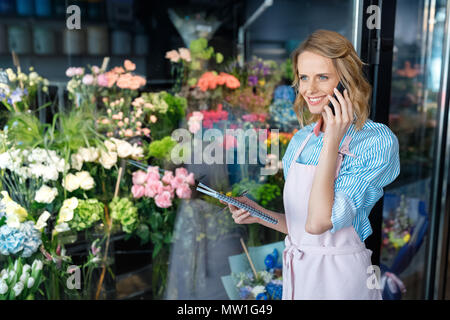 smiling young florist talking on smartphone while holding notebook in flower shop Stock Photo