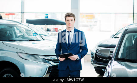 Handsome manager standing between cars in car showroom and looking at camera Stock Photo