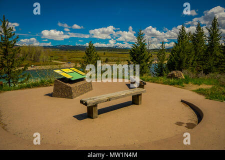 YELLOWSTONE, MONTANA, USA MAY 24, 2018: Beautiful outdoor view of informative sign and wooden public chair with a view of Jackson Lake Dam in Grand Teton National Park Stock Photo