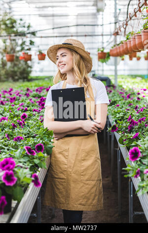 Attractive female gardener holding clipboard by flowers in greenhouse Stock Photo