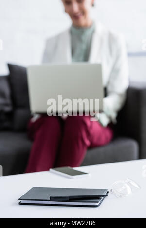 selective focus of businesswoman using laptop, smartphone, diary, pen and glasses lying on table on foreground Stock Photo