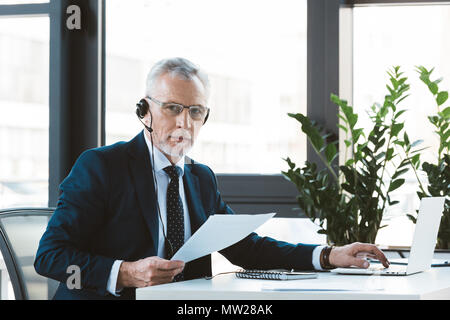 serious businessman in eyeglasses and headset using laptop and looking at camera Stock Photo