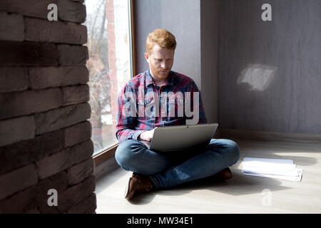 Young freelancer working from home. Attractive guy with ginger hair and beard sitting crosslegged with computer on his lap leaning against wall. Stock Photo