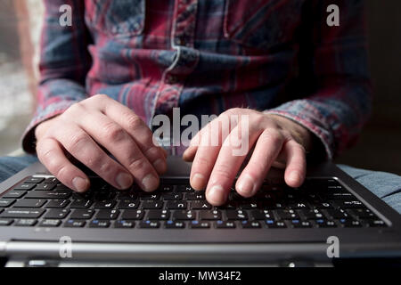 Mans hands typing on laptop keyboard. Front view of young man wearing jean shirt using computer. Stock Photo