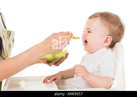 cropped image of mother in apron feeding crying baby boy in highchair isolated on white background Stock Photo
