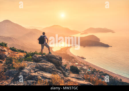 Young sporty man with backpack standing on the top of rock and looking at the seashore and mountains at sunset in summer. Scene with man, sea, mountai Stock Photo