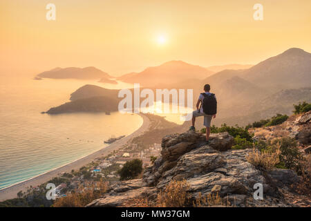 Young sporty man with backpack standing on the top of rock and looking at the seashore and mountains at sunset in summer. Scene with man, sea, mountai Stock Photo