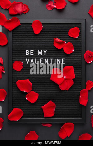 Black letterboard with white plastic letters with quote Be My Valentine, with red rose petals on black background. Stock Photo