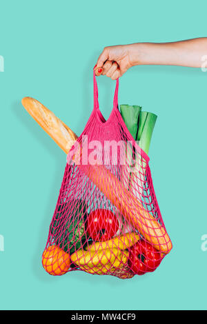 Woman hand holding a fuchsia string shopping bag with vegetables, fruits and bread in front of a green mint background Stock Photo