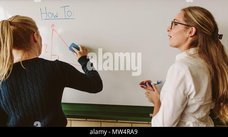 Rear view of young student writing on board with teacher standing by in classroom. Student writing on board during geometry class. Stock Photo