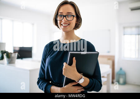 Smiling businesswoman wearing spectacles standing in office holding a diary. Portrait of a happy woman entrepreneur at work. Stock Photo