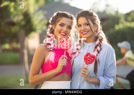Girlfriends with colorful braids holding candy heart on stick. Stock Photo