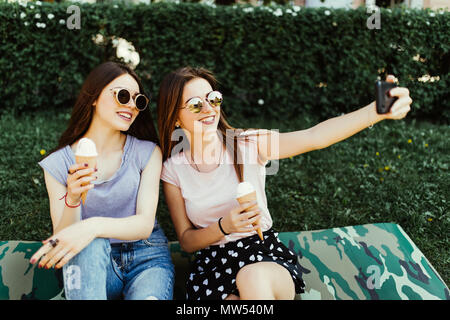 Portrait of two young women standing together eating ice cream and taking selfie photo on camera in summer street. Stock Photo