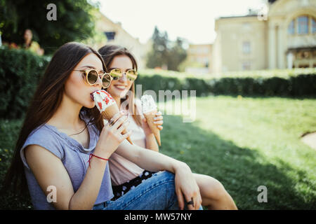 Young two women friends eat ice cream sitting on the grass in city streets