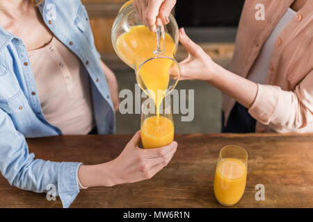 two women, senior and young drinking orange juice at table in kitchen. Woman pouring juice into glass Stock Photo