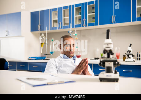 Young thoughtful technician in lab coat sitting in laboratory with microscope on table Stock Photo