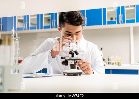 Young lab technician in white robe using microscope for sample analysis in laboratory Stock Photo