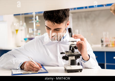 Young lab technician in white coat taking notes and using microscope in laboratory Stock Photo