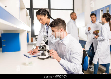 Young lab technicians working with microscope and taking notes on analysis on clipboard Stock Photo