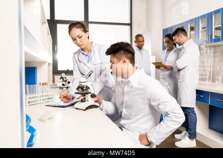 Young lab technicians working with microscope and taking notes about analysis on clipboard Stock Photo