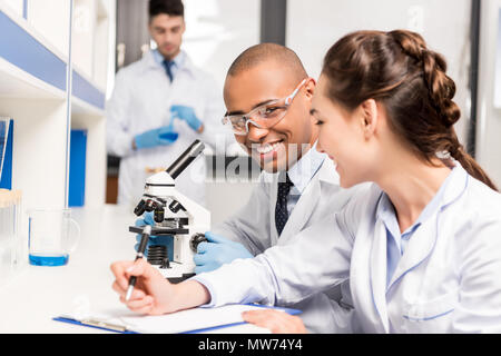 side view of smiling scientists working in laboratory with microscope and taking notes Stock Photo