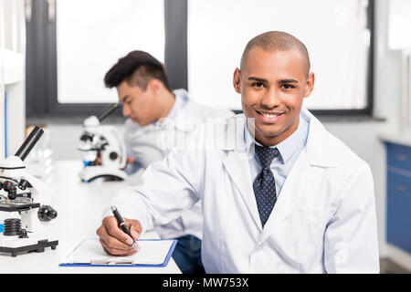 Young handsome doctor in lab coat writing notes on clipboard and smiling Stock Photo