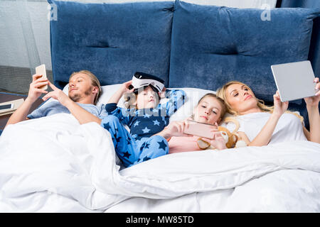 family with two children using digital devices while lying together in bed Stock Photo