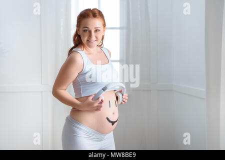 charming redhead pregnant woman with headphones on her belly sitting on  sofa with teddy bear, Stock image