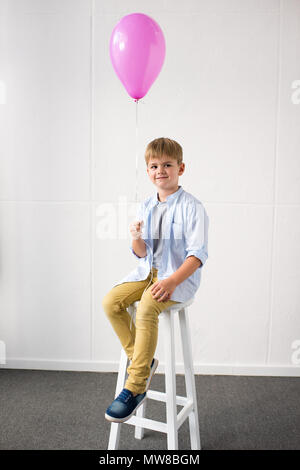 adorable little boy holding pink balloon while sitting in stool at birthday party Stock Photo