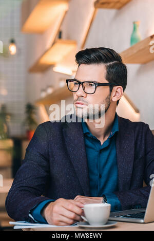 portrait of pensive businessman looking away while sitting at table with laptop in cafe Stock Photo