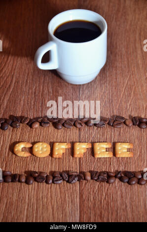 Still life with a small white cup of hot coffee and a salt cracker in the form of letters forming the word "Coffee", framed by rows of coffee beans on Stock Photo