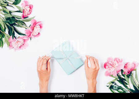 Woman opening her present, top view. Female's hands pull silver ribbon to unwrap blue gift box among the pink peony flowers, festive flat lay arrangem Stock Photo