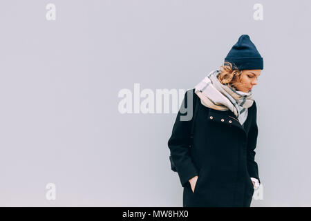 Young woman standing outdoors in cold weather on grey wall background. Female wearing coat, beanie hat and scarf, looking down, copy space. Stock Photo