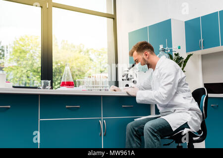 Concentrated young scientist in protective mask working with microscope in chemical lab Stock Photo