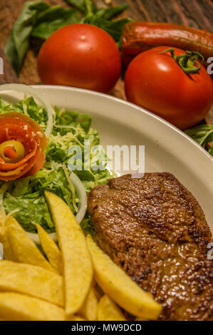 Close up of detailed well done grilled New York steak with french fried potatoes, tomatoes, sausage. served in white plate on cutting board on wooden table background Stock Photo