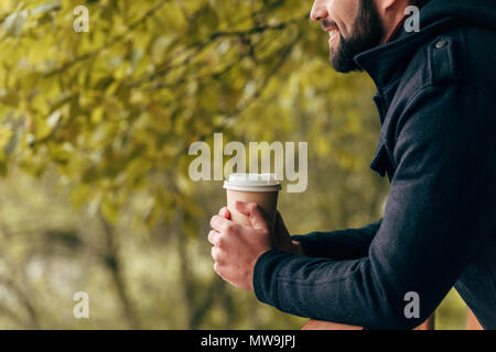 side view of smiling young man holding disposable coffee cup in autumn park Stock Photo